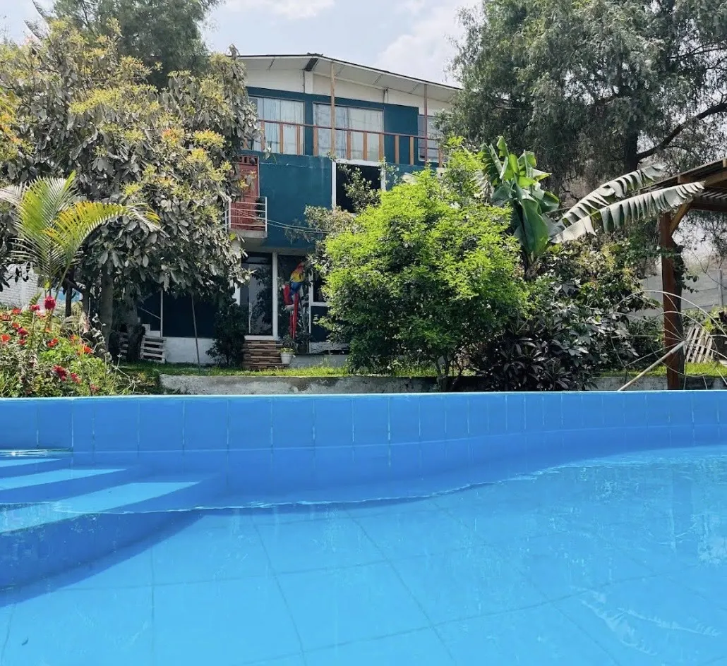 La casa de dos pisos color turquesa vista desde la piscina, rodeada de árboles